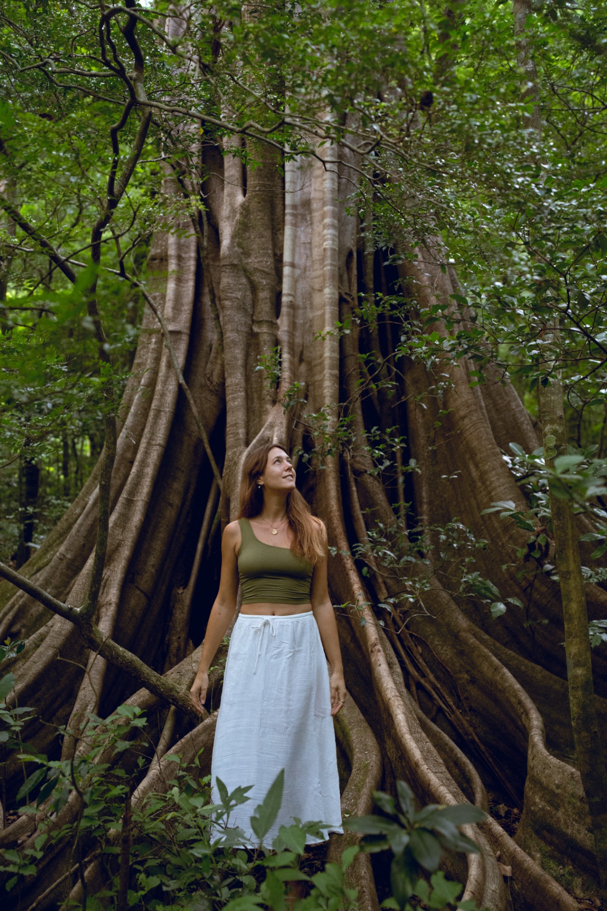 A woman standing peacefully at the base of an ancient tree, surrounded by lush forest.