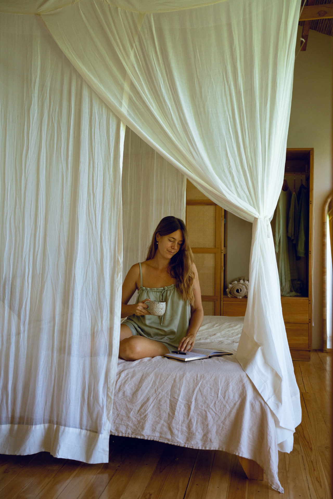 A woman journaling with a warm drink in a calm, sunlit bedroom.